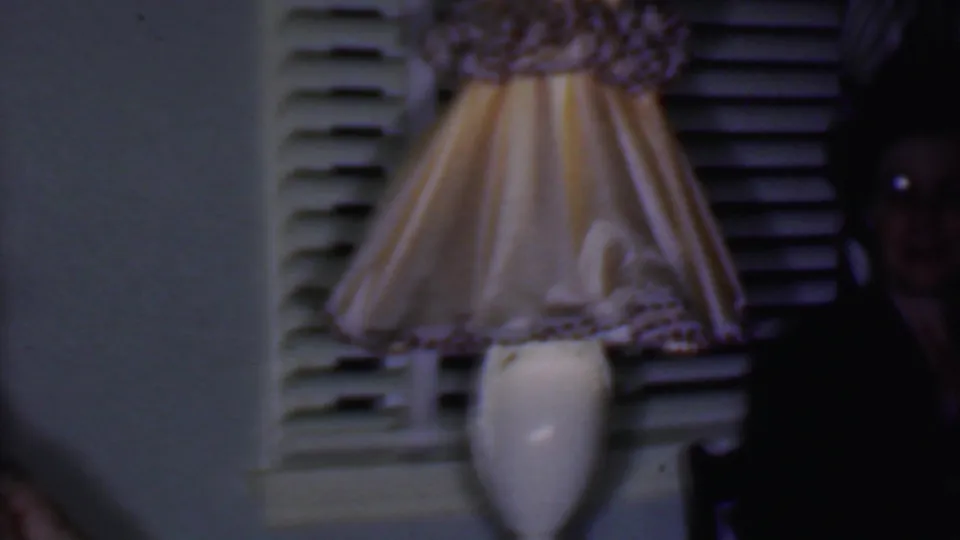 A table lamp with a patterned shade stands in the foreground of the 1956 Phoenix interior, with a partially visible adult at the right edge.