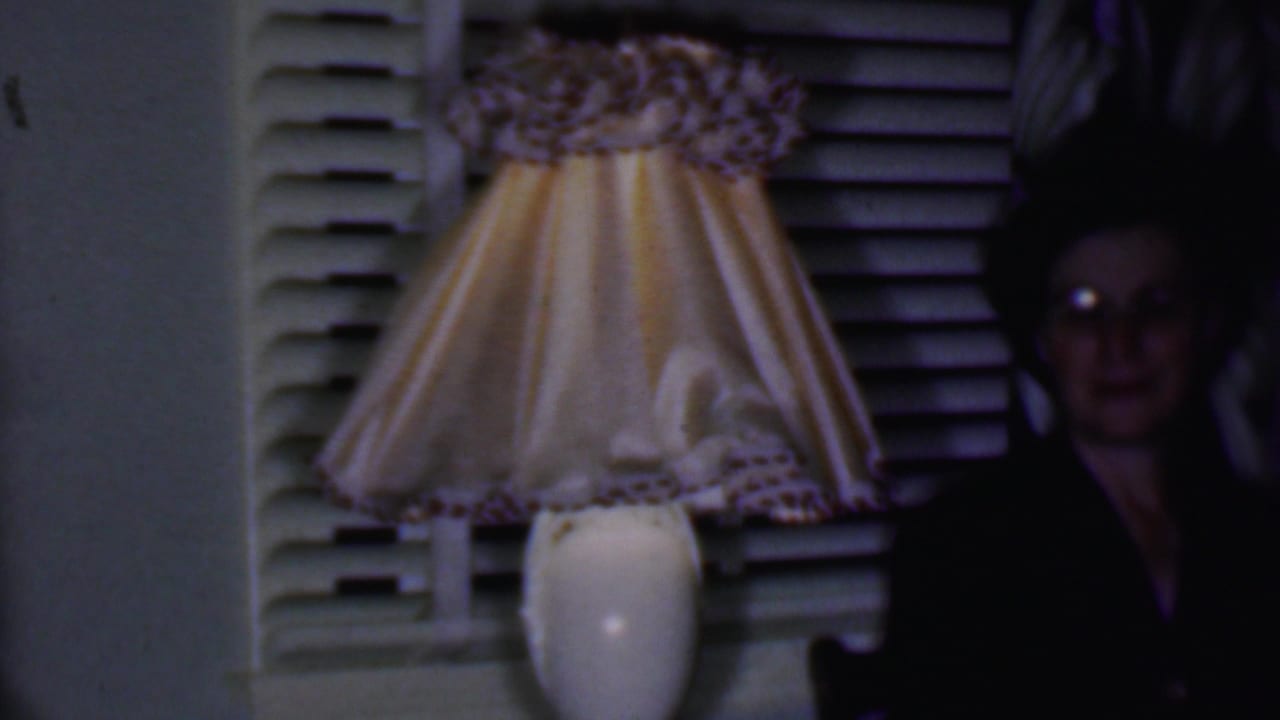 In a dim 1956 Phoenix home interior, a woman in dark clothing and glasses sits beside a glowing lamp with a pleated yellow shade in front of white blinds.