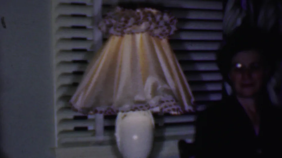 In a dim 1956 Phoenix home interior, a woman in dark clothing and glasses sits beside a glowing lamp with a pleated yellow shade in front of white blinds.