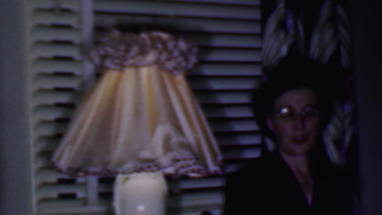 Woman in glasses near a lit pleated lamp, window blinds, and dark patterned background.
