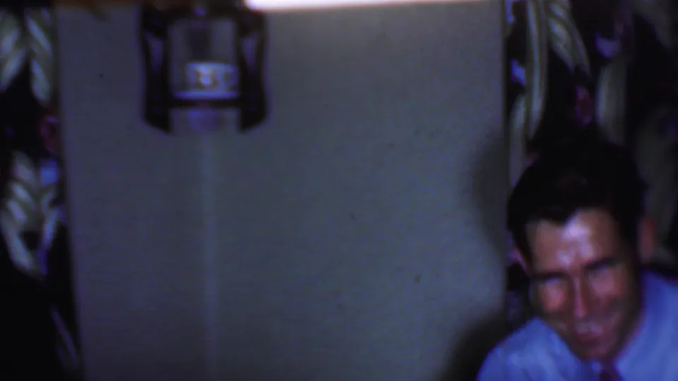 A man in a light shirt and tie smiles or laughs at the edge of the dim 1956 Phoenix bedroom scene, with wall decor and dark patterned background nearby.