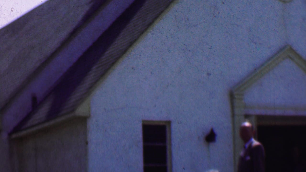 1956 Dallas home-movie frame showing a white stucco building exterior with a dark open doorway and formally dressed adults near the entrance.