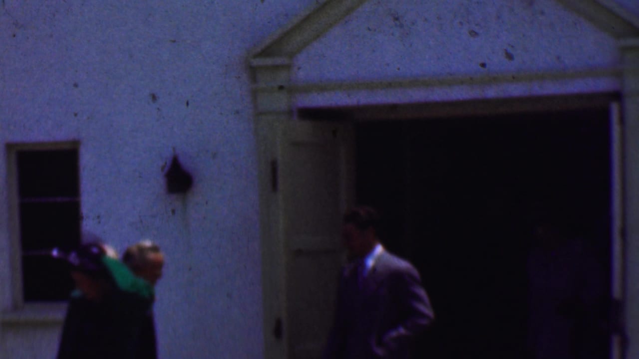 Several formally dressed adults stand or move beside an open doorway on a stucco building.