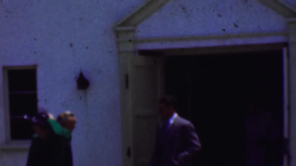 1956 Dallas 8mm frame with suited men and a woman in dark formal clothing near an open doorway in a light stucco exterior.