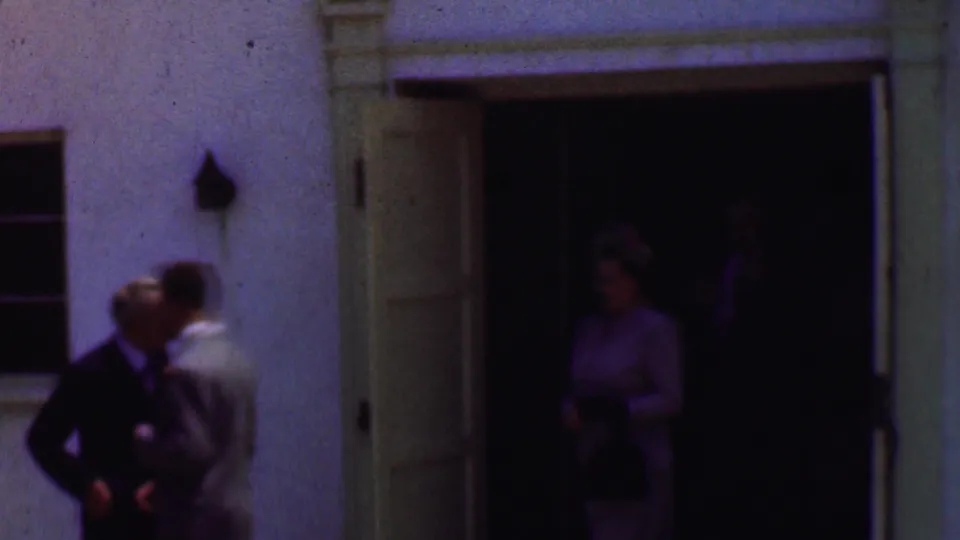 1956 Dallas home-movie still showing two suited men near the stucco wall while a woman stands partly visible inside the dark doorway.