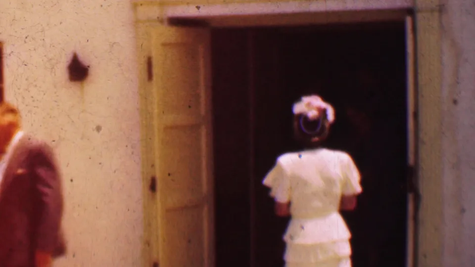 1956 Dallas 8mm frame showing a woman in a light formal dress and hat approaching or entering a dark open doorway on a stucco building.