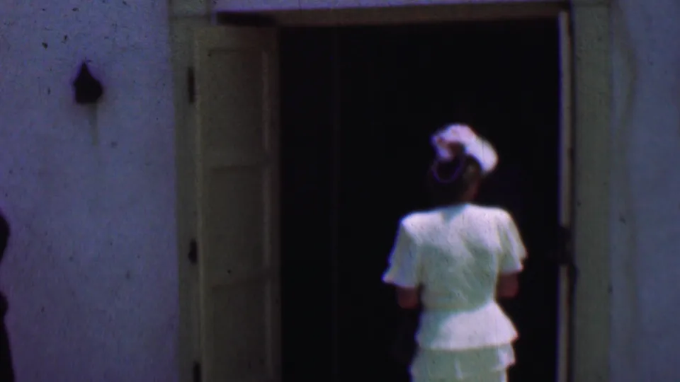 In 1956 Dallas home-movie footage, a woman in a white dress and hat approaches or pauses at the open doorway of a white church building.