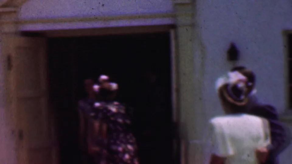 The final frame shows the woman in white and the suited man near the white church facade, while a floral-dressed woman remains partly visible inside the dark doorway.