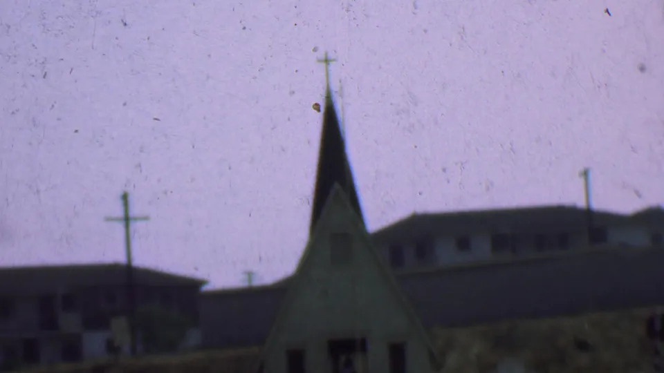 A small church steeple with a cross is seen in the supplied Dallas 1956 footage, with low background buildings and utility poles under bright sky.