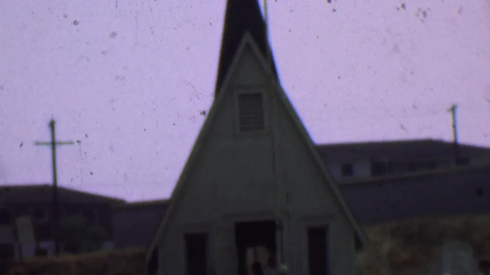 The camera frames the front of the small church, showing the steep roofline, cross-topped steeple, dark entrance, and nearby poles in faded 8mm color.