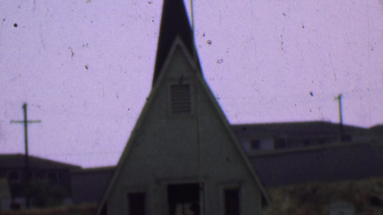 1956 Dallas home-movie frame showing a small white church exterior with a steep steeple, cross, utility pole, grass, and background buildings.