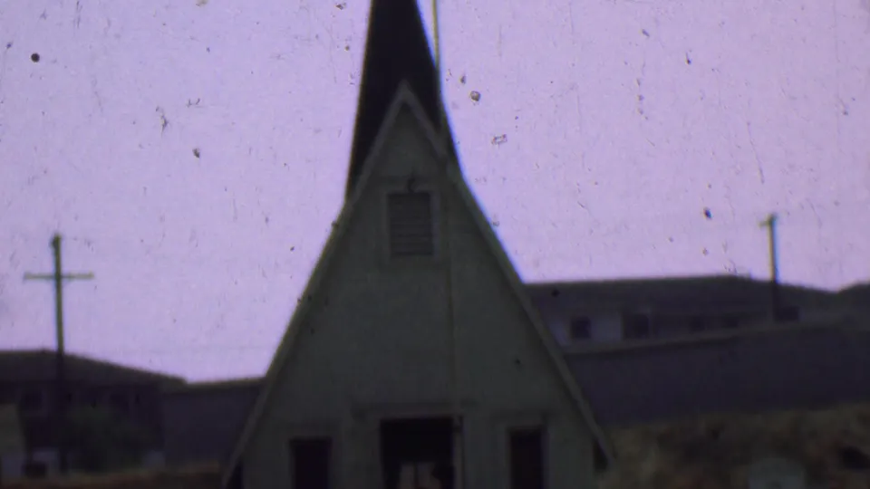 1956 Dallas home-movie frame showing a small white church exterior with a steep steeple, cross, utility pole, grass, and background buildings.