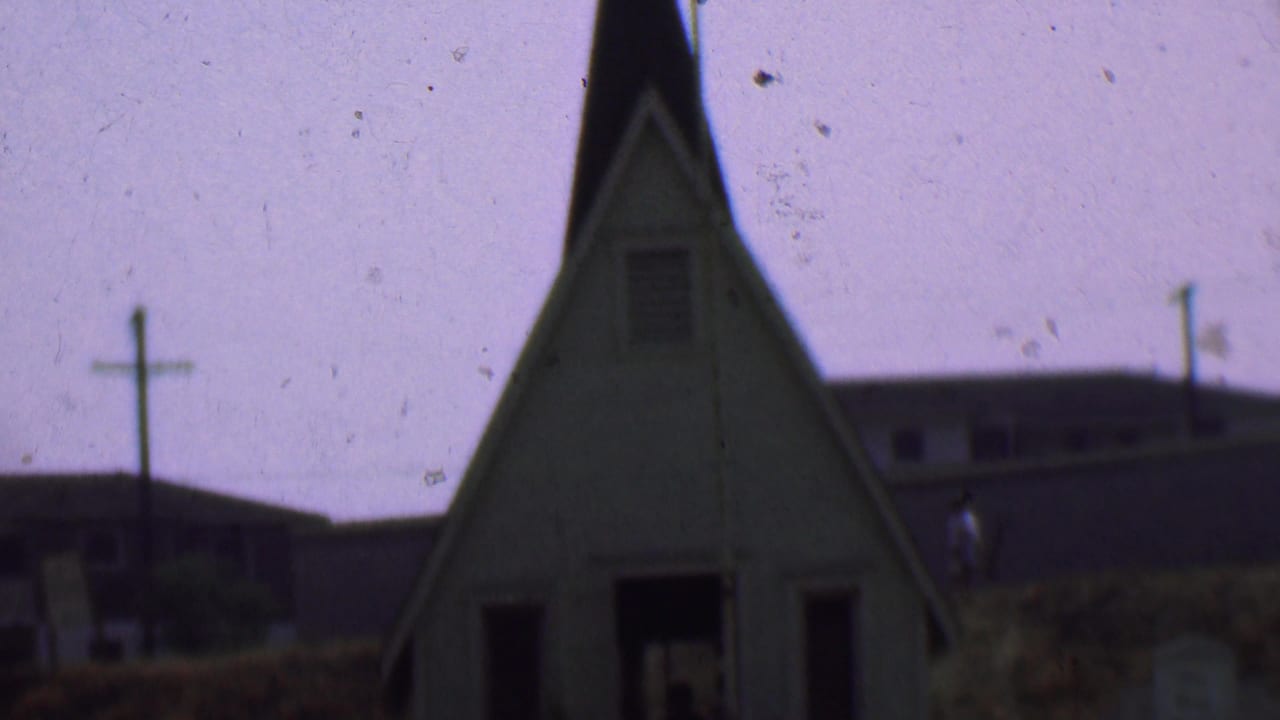 1956 Dallas frame showing the upper portion of a small church with cross, steep roofline, utility poles, and a low building in the background.