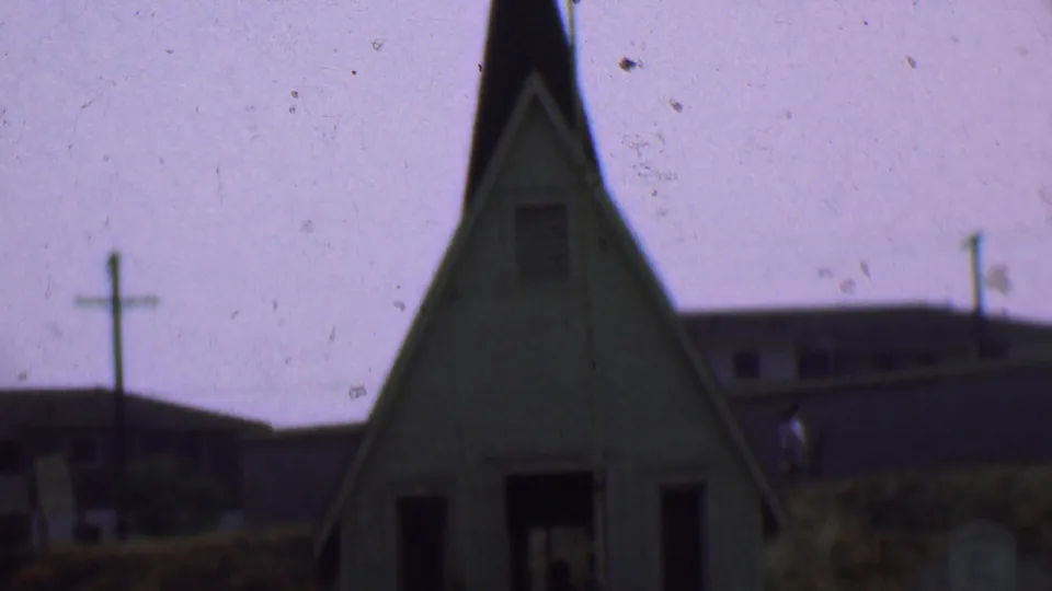 1956 Dallas frame showing the upper portion of a small church with cross, steep roofline, utility poles, and a low building in the background.