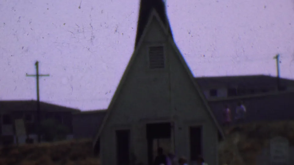 1956 Dallas home-movie frame showing a small white church exterior with people near the dark entrance, a cross, grass, utility poles, and background buildings.