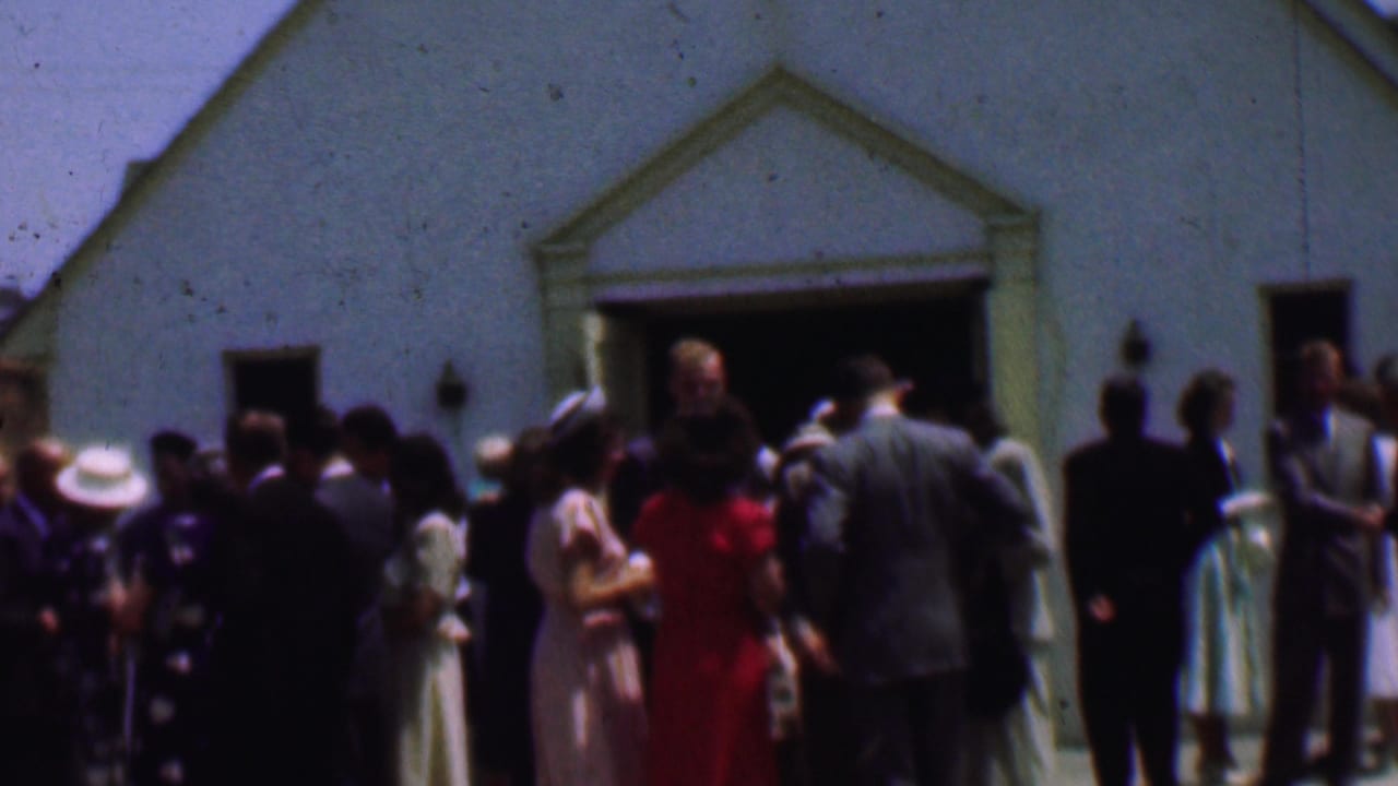 Dallas 1956 home-movie frame showing adults in suits, dresses, and hats gathered outside a white church after services.