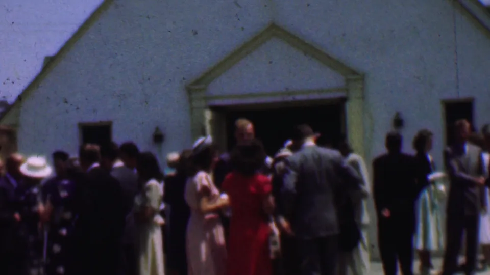 Dallas 1956 home-movie frame showing adults in suits, dresses, and hats gathered outside a white church after services.