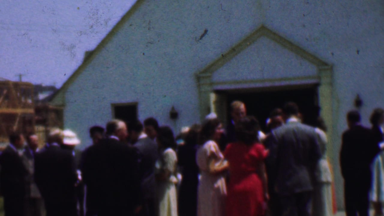 Dallas 1956 frame of adults socializing outside a white church, with a wood-frame structure under construction in the background.