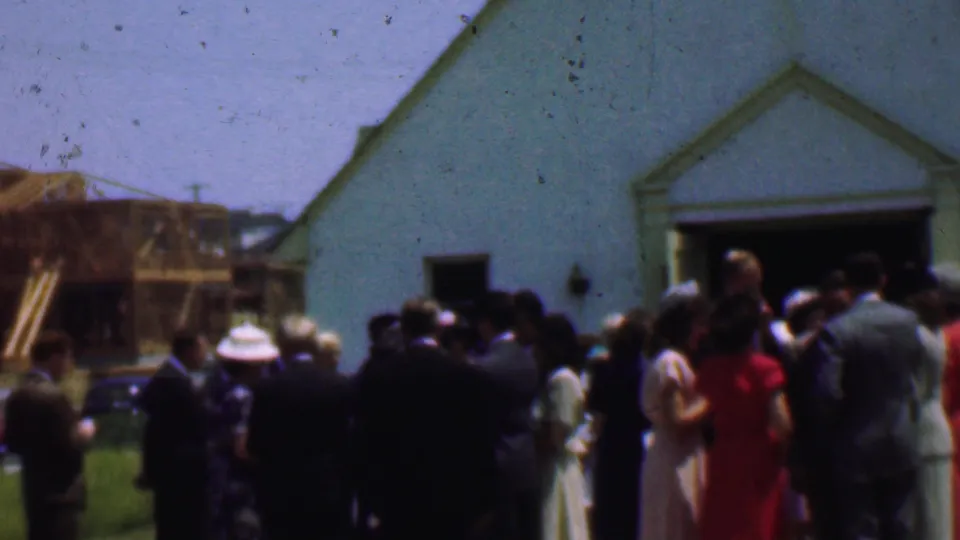 Dallas 1956 home-movie frame looking across a crowd in formal Sunday clothing toward a white church and nearby wood construction.