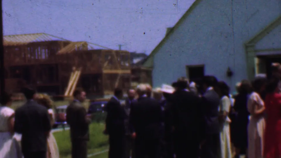 Dallas 1956 frame showing adults gathered beside a white church, with parked cars, grass, and a wooden building frame in the background.