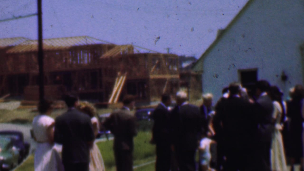 Dallas, Texas, 1956 home-movie frame showing a small group gathered outside a church on a sunny lawn, with parked cars and a partially framed wooden building behind them.