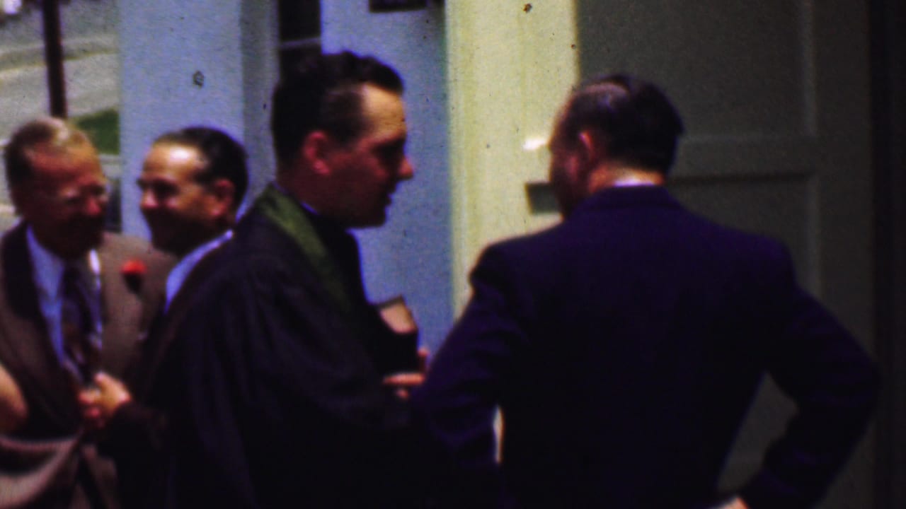 Men in suits and a clergyman stand near a church entrance talking.