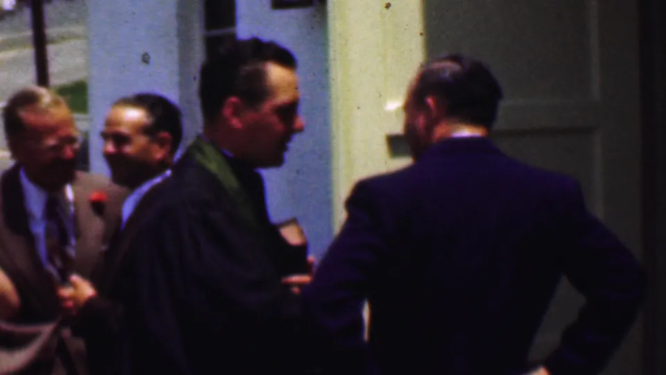 Dallas, Texas, 1956 frame showing suited men and a robed clergyman with a green stole conversing near a light-colored church entrance.