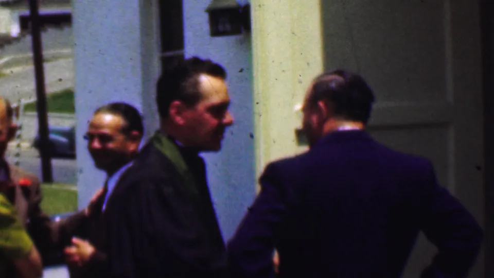 Dallas, Texas, 1956 home-movie still showing a robed clergyman and a suited man beside a church entrance, with another attendee partly visible at the edge of the frame.