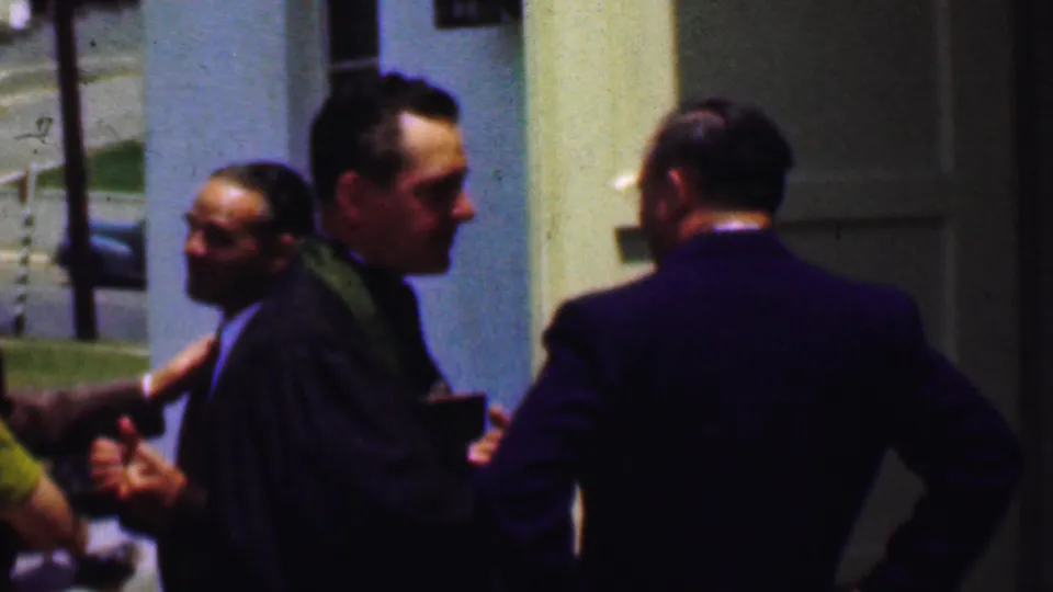 Dallas, Texas, 1956 frame showing a robed clergyman near a church entrance with suited men and other attendees gathered close by in bright daylight.