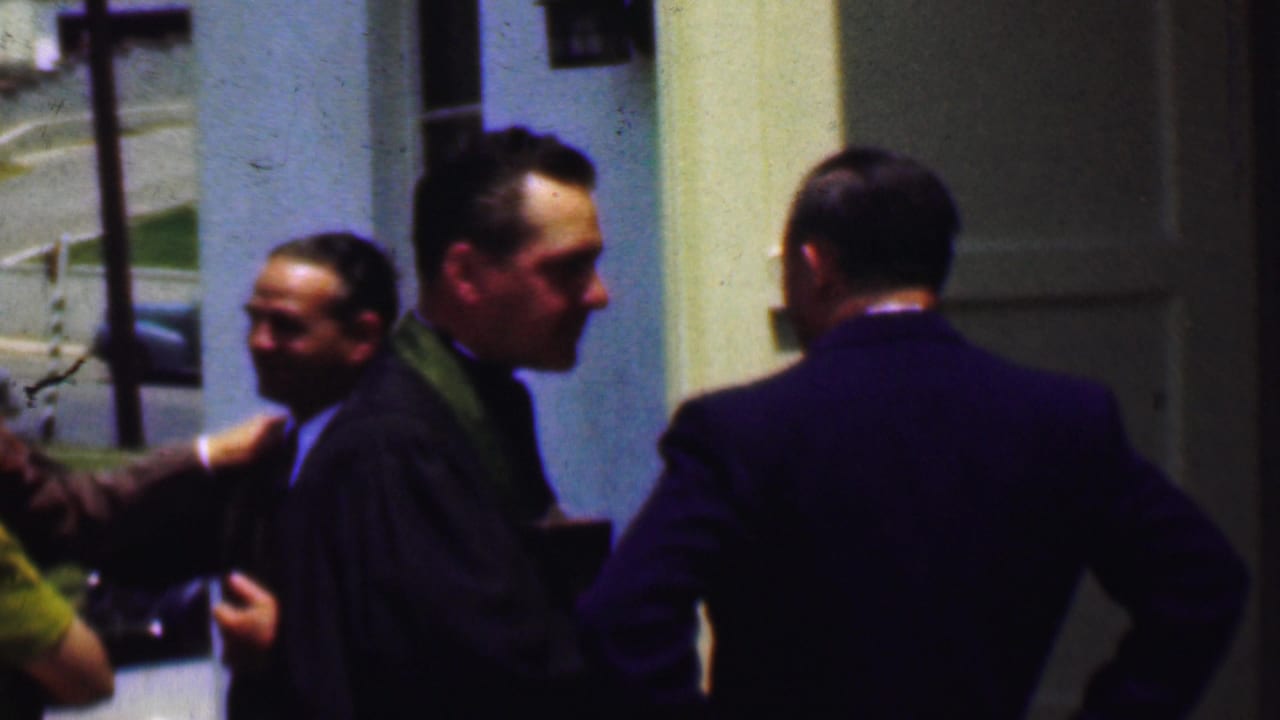 In 1956 Dallas home-movie footage, formally dressed adults stand outside a church entrance while a clergyman in a green stole speaks near parked mid-century cars.