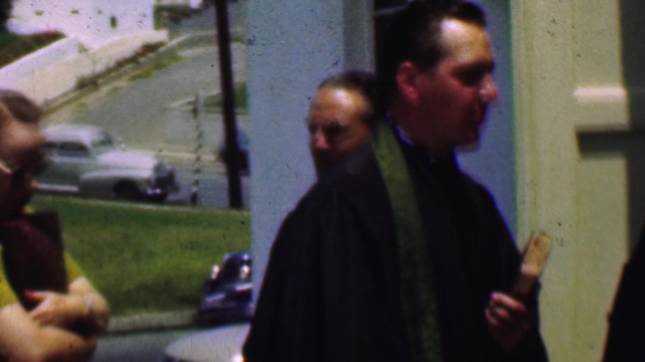 A clergyman in dark clothing and a green stole holds a small book outside a Dallas church, with a pale building facade and street scene behind him.