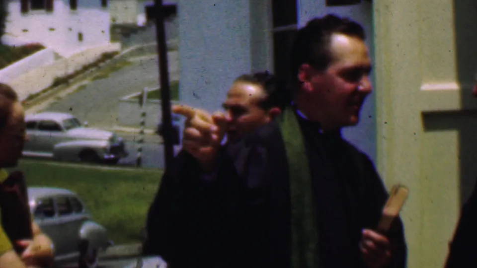 A clergyman in green stole points during conversation outside a Dallas church, with adults, a pale facade, parked cars, and a lawn visible in the 8mm frame.