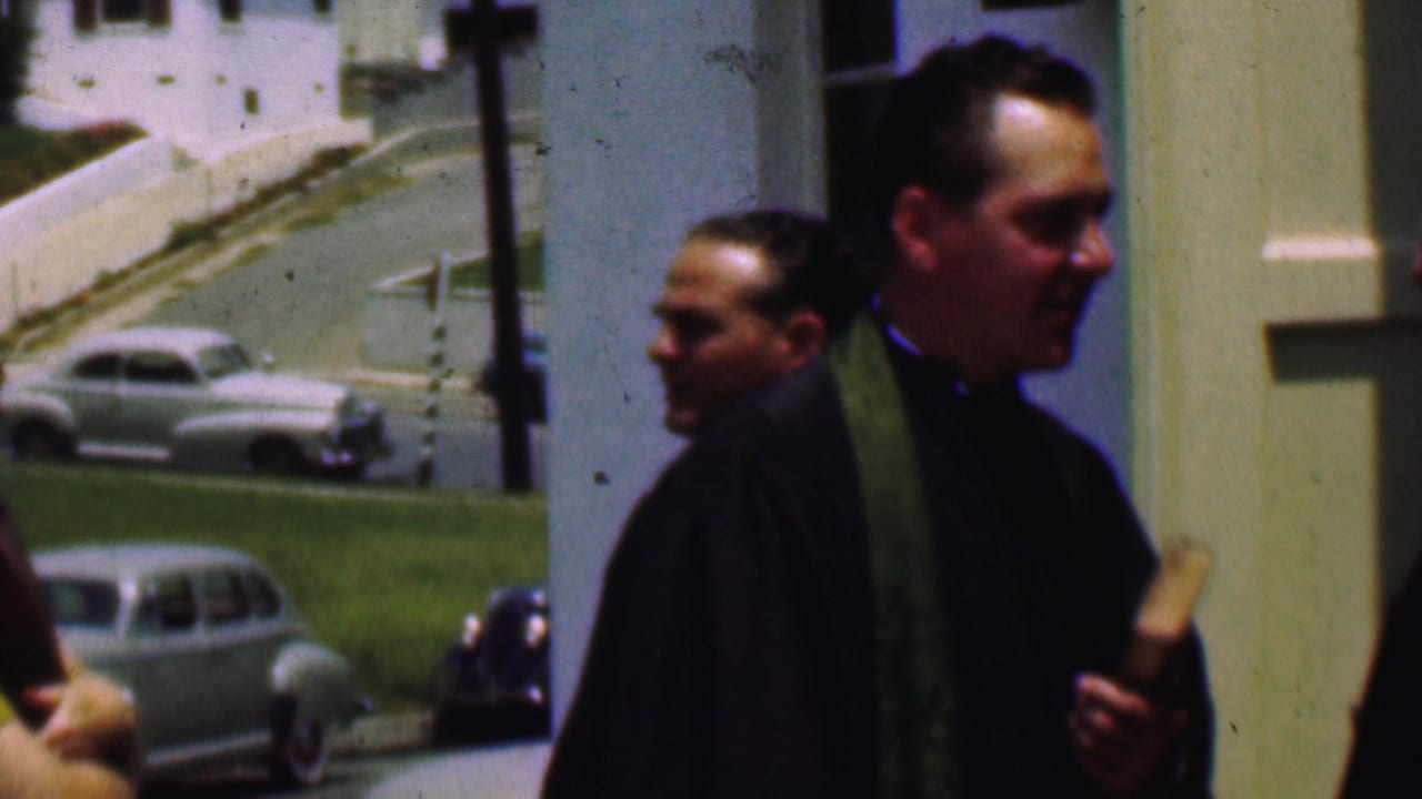 In 1956 Dallas, a priest in dark clerical clothing and a green stole walks near a church entrance while mid-century cars sit along a grassy roadside behind him.