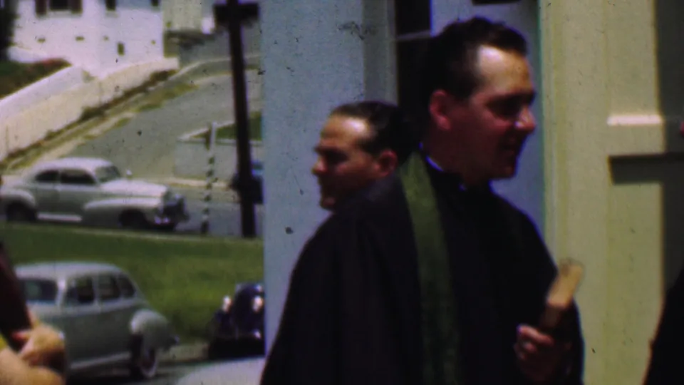 In 1956 Dallas, a priest in dark clerical clothing and a green stole walks near a church entrance while mid-century cars sit along a grassy roadside behind him.