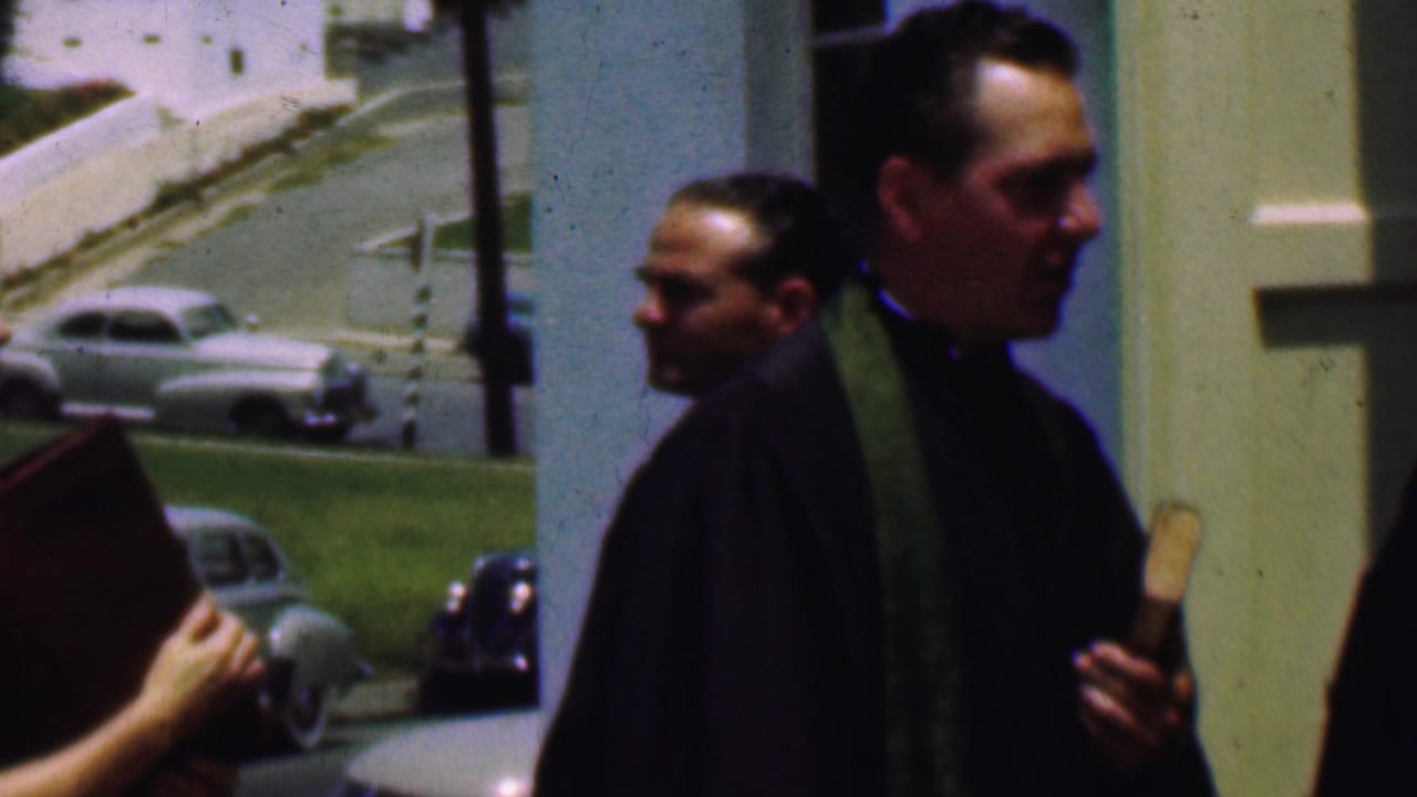 Priest and parishioners pass near a church entrance with parked cars in the background.