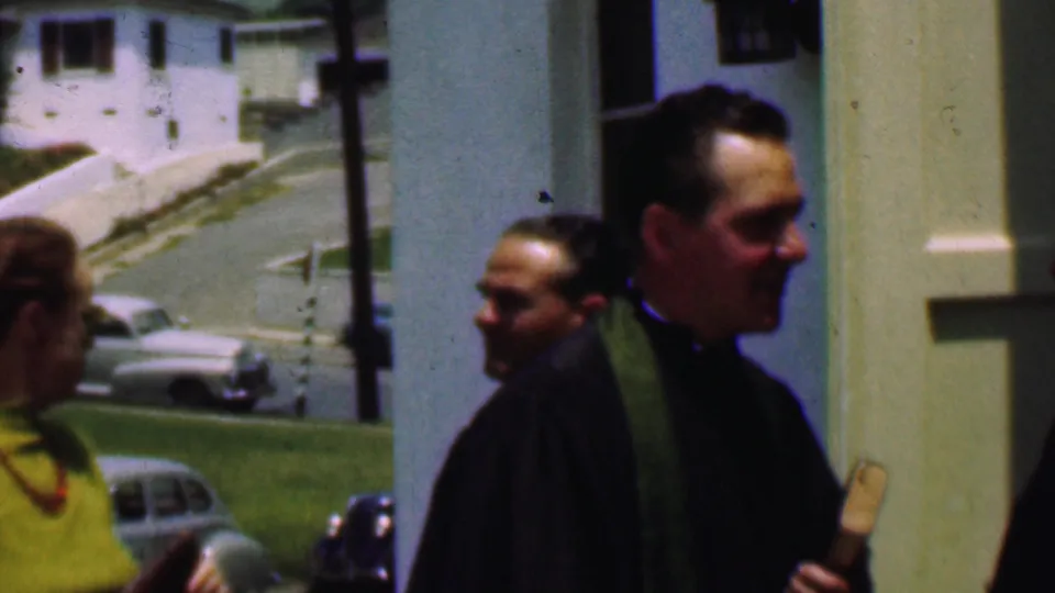 The handheld 8mm frame shows the priest and parishioners moving outdoors near church walls, with rounded mid-century cars parked along the roadside in Dallas.