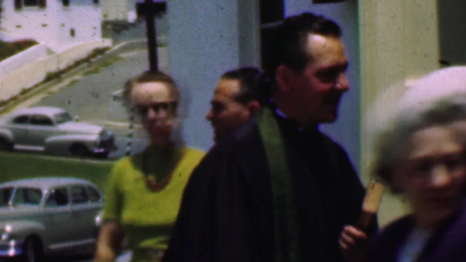 In a crowded outdoor church moment in 1956 Dallas, a priest stands near passing parishioners while a woman in yellow and parked period cars appear behind him.
