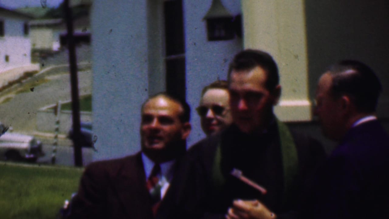 In a Dallas 1956 home-movie frame, a priest wearing a green stole stands outside a church with suited adults and a woman in sunglasses nearby.