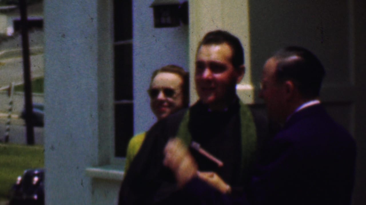 The priest, wearing a green stole, holds a small book or object while speaking with a suited man outside the Dallas church setting described in the clip metadata.