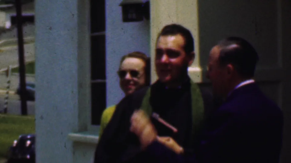 The priest, wearing a green stole, holds a small book or object while speaking with a suited man outside the Dallas church setting described in the clip metadata.