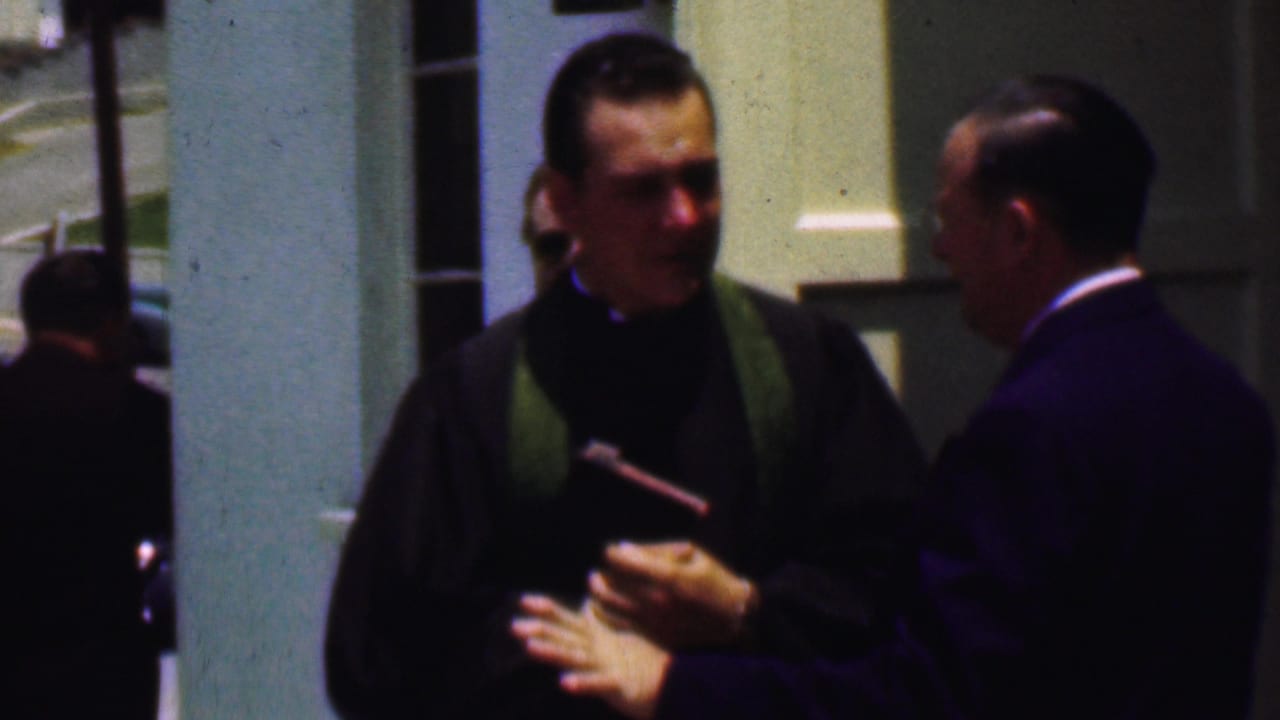 A Dallas 1956 frame shows the priest and suited man in close conversation outside the church, with pale columns, lawn, and automobiles visible behind them.