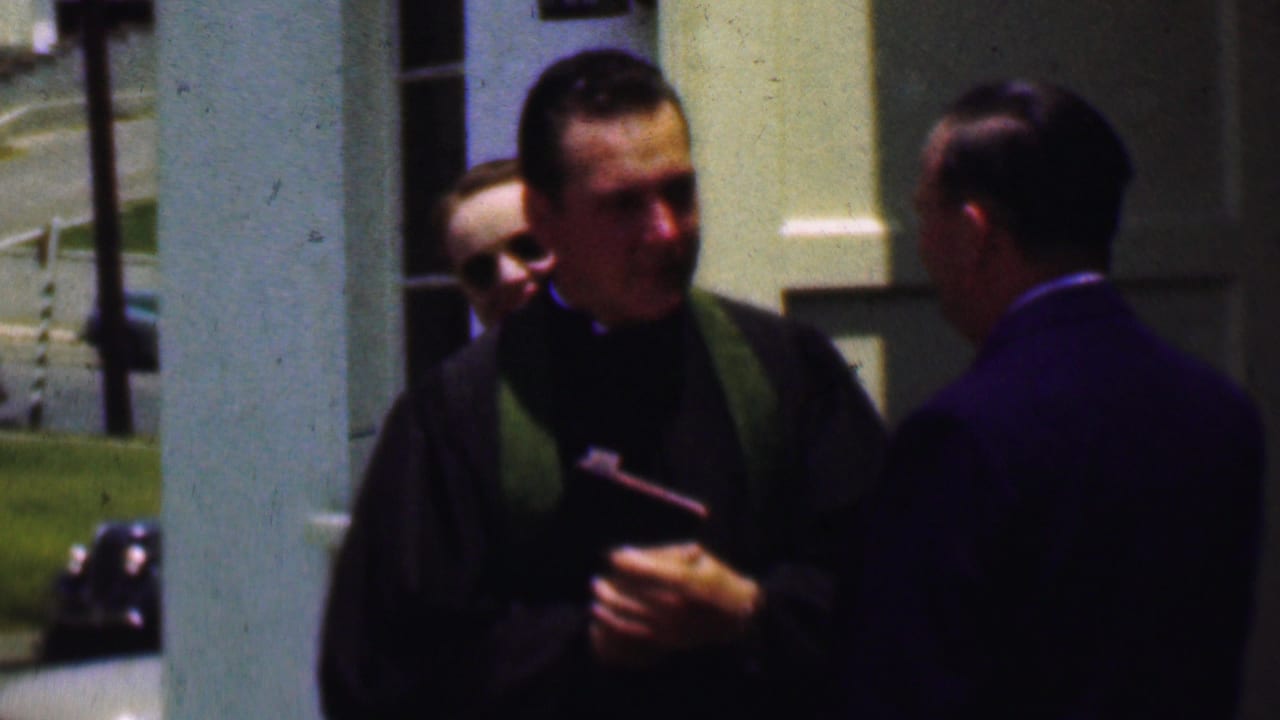 The closing frame keeps the priest and suited man in profile outside the church, with another adult partly visible between them and the columns in the background.