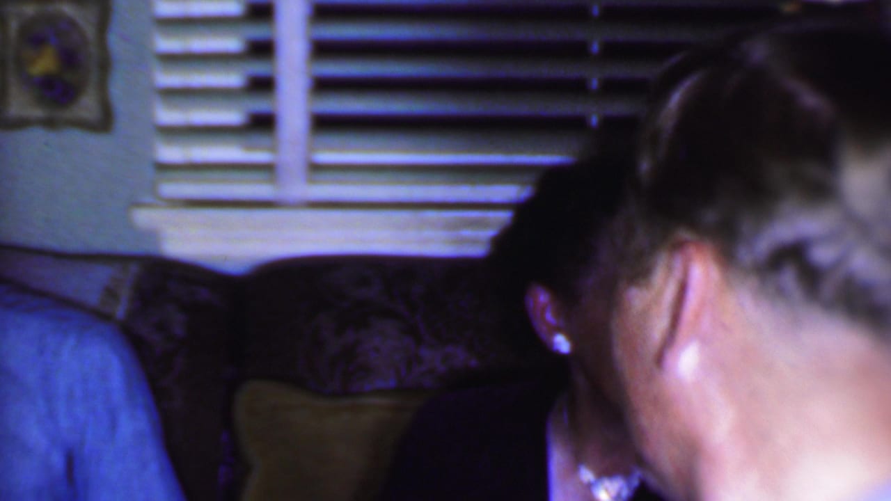 1956 Phoenix home-movie frame showing adults seated close together in a dim private living room with horizontal blinds and upholstered furniture behind them.