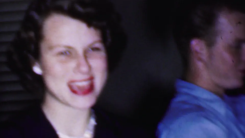 1956 Phoenix home-movie frame with a smiling woman wearing pearls and dark clothing seated beside a man in a light blue collared shirt inside a private room.