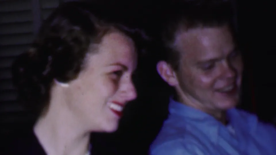 1956 Phoenix home-movie frame showing a woman in profile smiling toward a seated man, with another man partially visible in the dark living room setting.