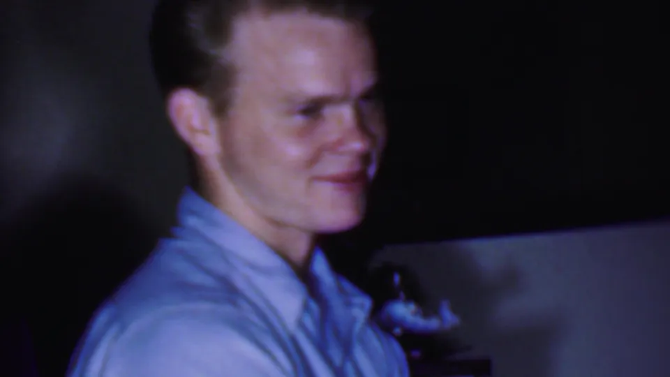 1956 Phoenix home-movie frame showing a seated man in a light blue collared shirt smiling in a dim private living room, with another adult partly visible behind him.