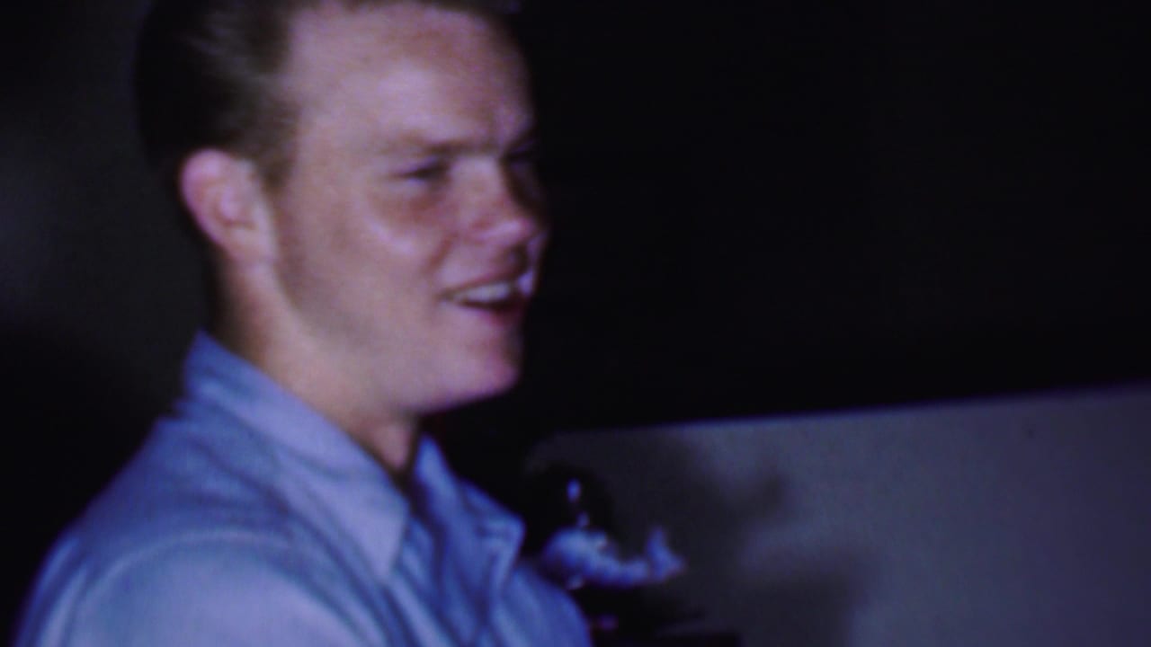 In 1956 Phoenix home-movie footage, a man in a light blue collared shirt smiles while standing or leaning in a dim private interior.