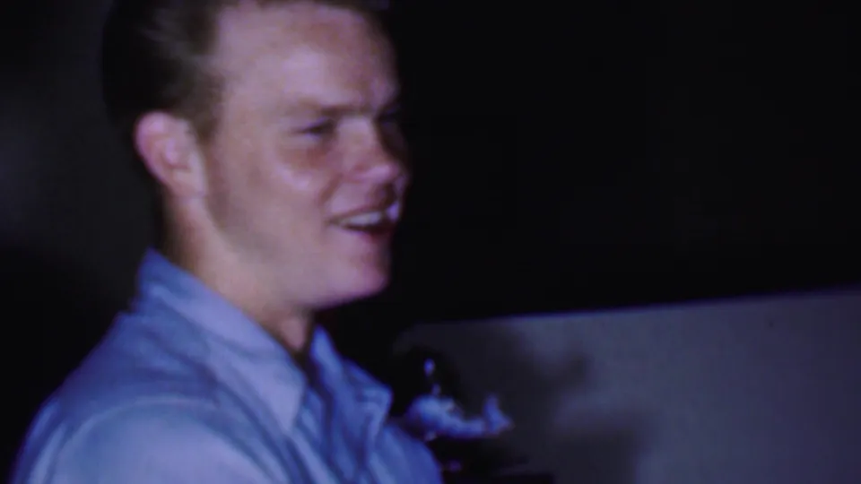 In 1956 Phoenix home-movie footage, a man in a light blue collared shirt smiles while standing or leaning in a dim private interior.