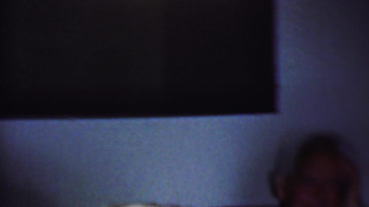 A shadowed seated man appears at the side of a dim private room, with a pale wall and dark window or wall opening behind him.