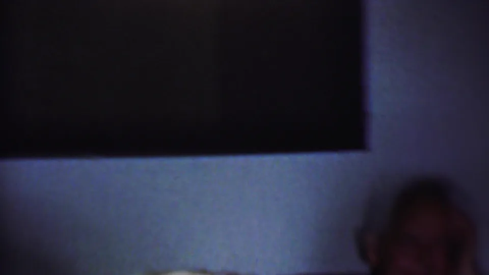A shadowed seated man appears at the side of a dim private room, with a pale wall and dark window or wall opening behind him.
